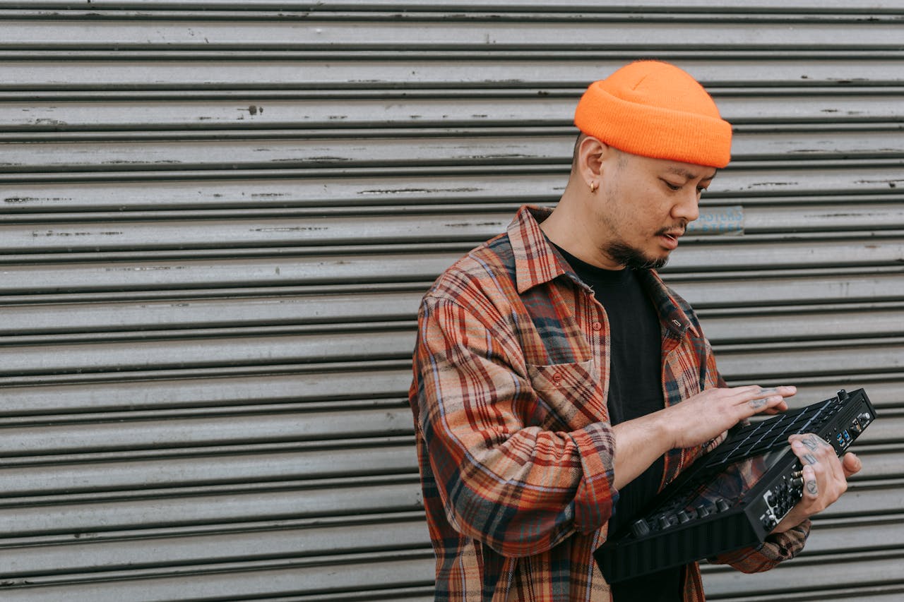 An adult man with an orange hat using an electronic mixer outdoors against a metallic background.